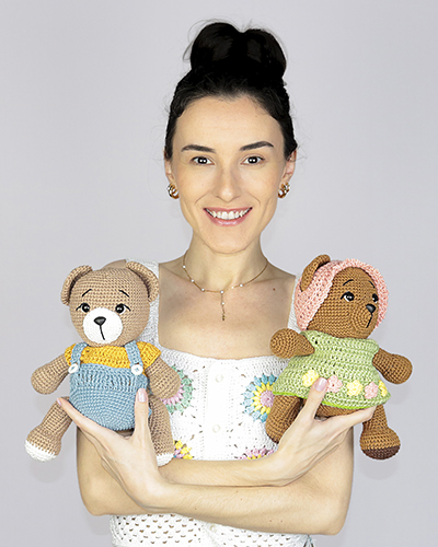 Foto de uma mulher branca com cabelos pretos de pé, segurando dois ursinhos feitos em amigurumi. O primeiro, veste blusa mostarda e jardineira azul. O segundo, vestido verde florido e chapéu rosa. A foto divulga um curso de amigurumi para iniciantes.