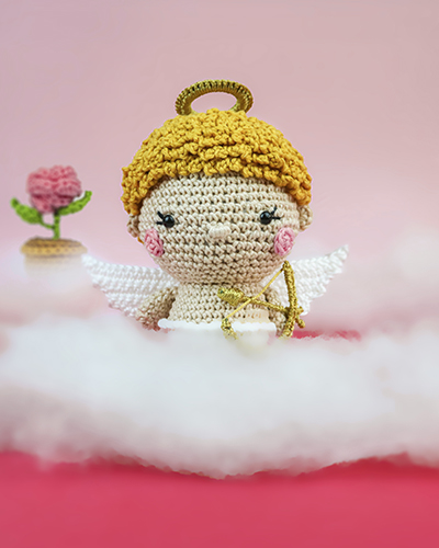 A foto mostra um bonequinho feito em amigurumi. É um anjo e cabelos enrolados loiros, auréola dourada e segura um arco e flecha. Veste somente um shortinho branco, que combina com suas asas, também brancas. O fundo é rosa, entre nuvens e pode-se ver uma flor rosa ao longe.